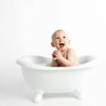 Cute baby laughing and clapping in a white bathtub against a white background.